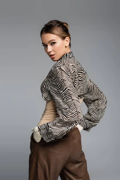 trendy woman in blouse with animal print, gloves and brown pants posing with hands in pockets isolated on grey