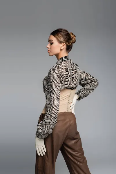 trendy woman in blouse with animal print, gloves and brown pants posing with hand on hip isolated on grey