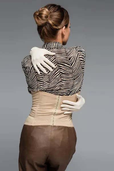 back view of woman in blouse with animal print, gloves and corset embracing herself isolated on grey