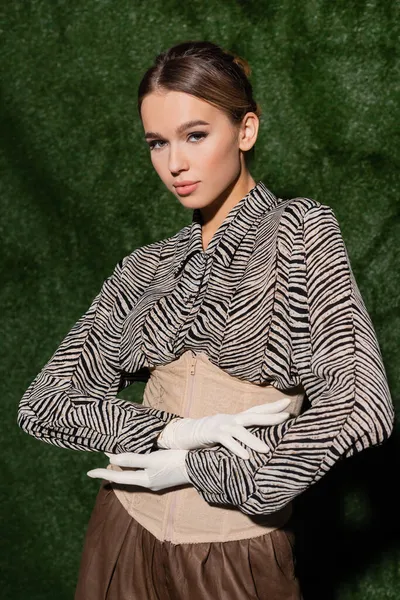 young trendy model in blouse with zebra print, corset and gloves looking at camera near grassy background