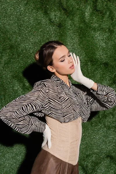 young trendy model in blouse with zebra print, corset and gloves posing with hand on hip near grassy background