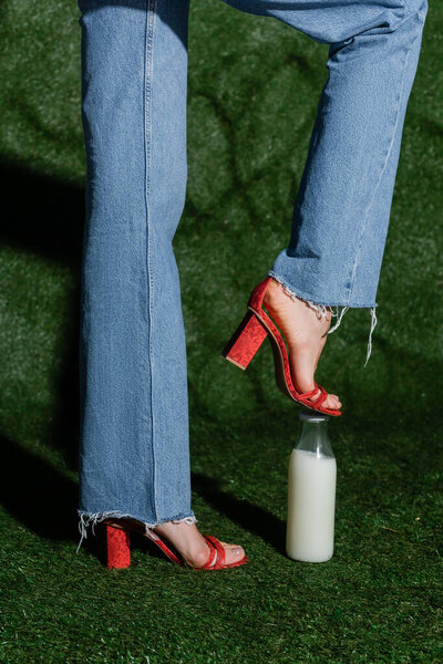 cropped view of stylish model in red high heels and jeans stepping on glass bottle of milk near grassy background 