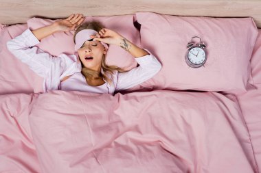 Top view of young woman in sleeping mask yawning near alarm clock on bed  clipart