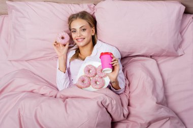 Top view of smiling woman holding donut and paper cup on bed at home  clipart