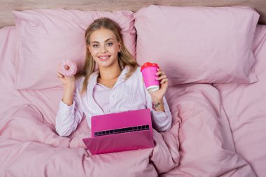 Pijamalı, elinde kağıt bardak ve laptopla yatağın yanında duran neşeli kadının en iyi görüntüsü. 