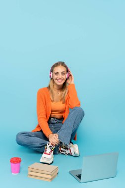 Happy blonde student in headphones sitting near laptop, paper cup and books on blue background  clipart