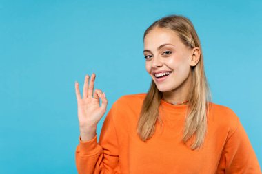 Cheerful woman in orange sweatshirt showing ok gesture isolated on blue  clipart