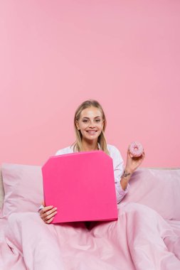 Pijamalı, gülümseyen sarışın kadın. Elinde çörek ve pizza kutusu var. Pembe yatakta tek başına. 