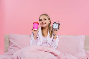 Pijamalı, elinde çalar saat ve kahve olan pozitif sarışın bir kadın pembe üzerine izole edilmek için bekliyor. 