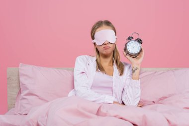 Uyku maskeli üzgün kadın, elinde çalar saatle pembe yatakta yalnız başına. 