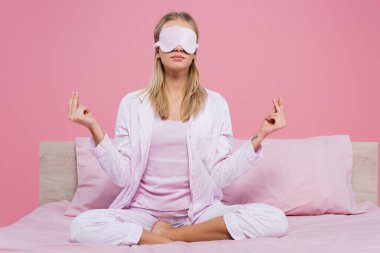 Uyku maskeli ve pijamalı genç bir kadın pembe yatakta meditasyon yapıyor. 