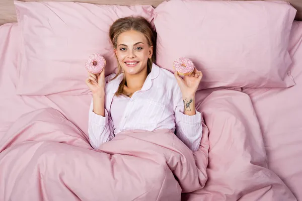Top view of positive blonde woman holding donuts and looking at camera on bed