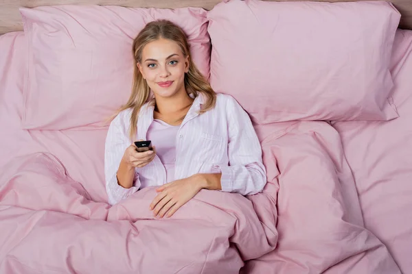 Top view of smiling blonde woman holding remote controller on pink bed