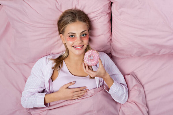 Top view of happy young woman with eye patches holding donut on bed 
