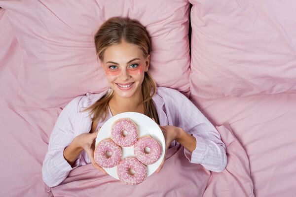 Top view of smiling woman with eye patches holding plate with donuts on bed 