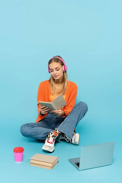 Blondynka Słuchawkach Czyta Książkę Pobliżu Papierowej Filiżanki Laptopa Niebieskim Tle — Zdjęcie stockowe