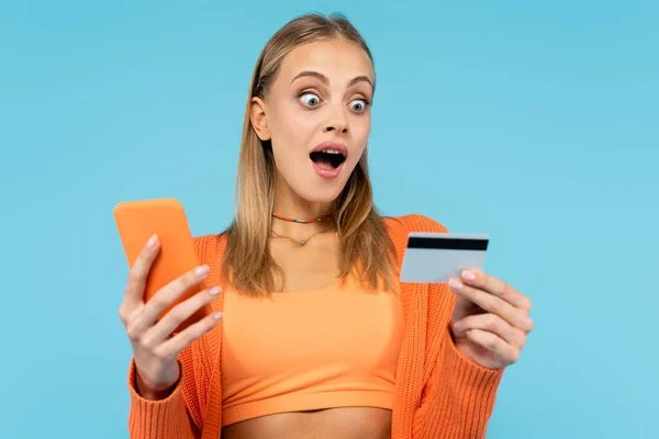 兴奋的金发女人带着信用卡和智能手机被蓝色隔离 — 图库照片