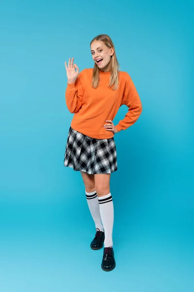 Full length of smiling student in skirt showing okay at camera on blue background