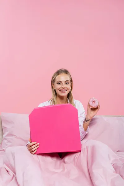 穿着睡衣 拿着甜甜圈和披萨盒躺在床上 与粉色隔离的金发女人笑着 — 图库照片