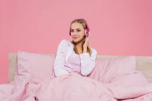 穿着睡衣的金发年轻女子在床上用耳机听着音乐 与粉色隔离 — 图库照片
