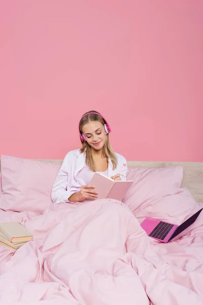 在笔记本电脑旁边的笔记本上写着的耳机里的自由职业者 在床上用粉色隔离的书 — 图库照片