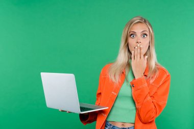 nervous blonde woman covering mouth with hand while holding laptop isolated on green clipart