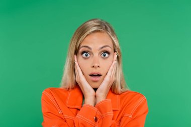 Şok olmuş turuncu ceketli kadın yeşil kameraya bakarken yüzüne dokunuyor.