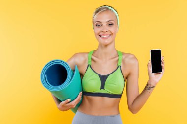 Yeşil spor sutyen giyen mutlu bir kadın spor minderi tutarken cep telefonunu sarı ekranda izole bir şekilde gösteriyor.