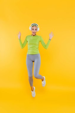 full length view of excited and sportive woman in headphones looking at camera while jumping on yellow clipart