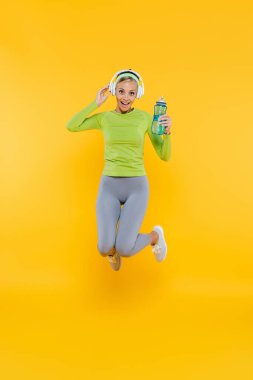 happy sportive woman in headphones jumping with sports bottle on yellow clipart