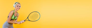 Sarı pankartta izole edilmiş tenis oynayan kulaklıklı, gülümseyen bir sporcu kadın.