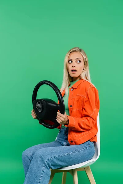 scared woman looking away while sitting on chair with steering wheel isolated on green