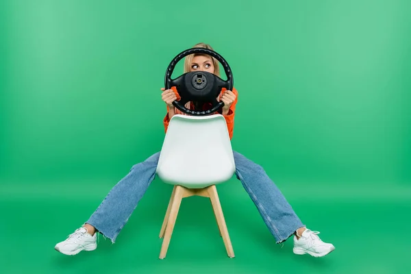 young woman in jeans obscuring face with steering wheel while sitting on white chair on green