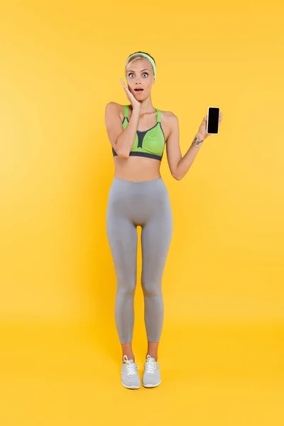 全景惊艳女子在运动服触摸脸同时显示智能手机与空白屏幕上的黄色 — 图库照片