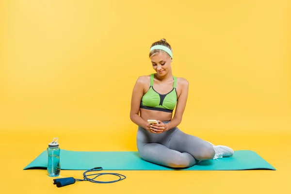 穿着运动服的女人坐在运动瓶旁边的健身垫上 坐在黄色的跳绳上 一边用智能手机高兴地看着 — 图库照片