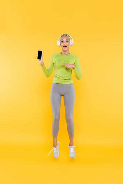 полный вид изумленной спортсменки в наушниках, указывающих на сотовый телефон с экраном банка на желтом