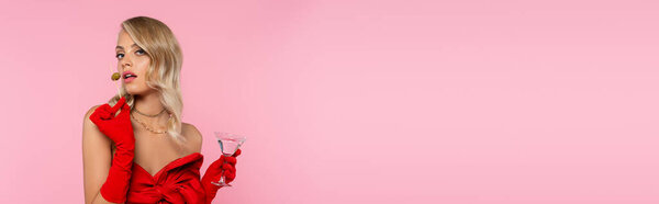 sensual blonde woman in red gloves holding drink and olives on cocktail skewer while posing isolated on pink, banner