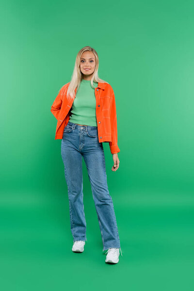 full length view of blonde woman in jeans and orange jacket standing with hand on hip on green