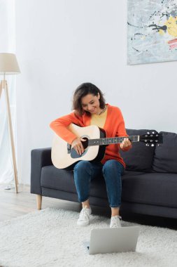 Halıda dizüstü bilgisayarın yanında akustik gitar çalmayı öğrenen neşeli genç bir kadın.