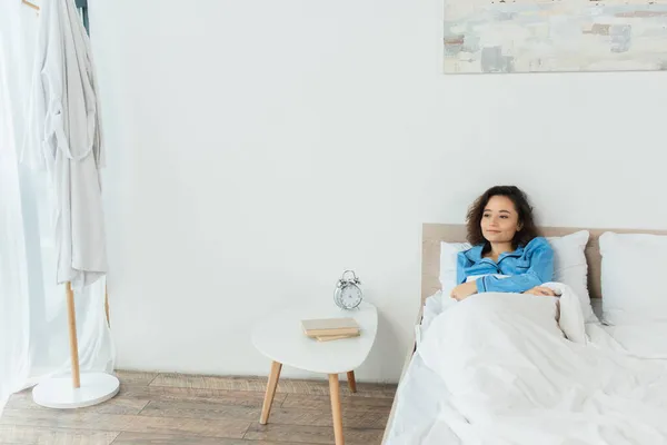 Pleased Woman Lying Bed Bedside Table Books Alarm Clock — Stock Photo, Image