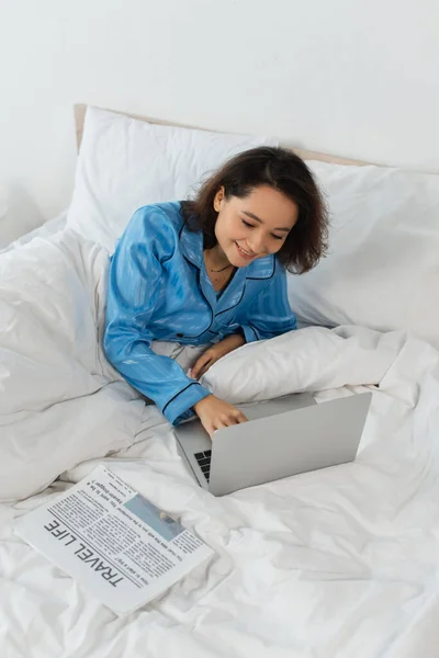 穿着睡衣的年轻自由职业者在床边用笔记本电脑微笑 — 图库照片