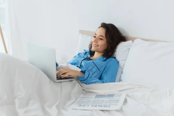 穿着蓝色睡衣的快乐的年轻自由职业者在床上靠报纸边使用笔记本电脑 — 图库照片