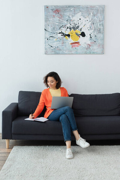 smiling woman writing in notebook while using laptop in living room 