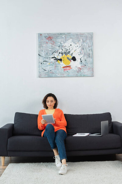 young woman holding digital tablet and sitting near laptop and notebook on couch 