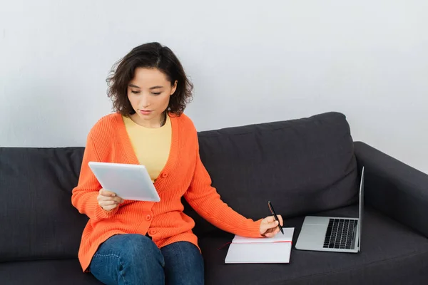 若い女性がデジタルタブレットを見てノートパソコンのそばでソファに書き — ストック写真
