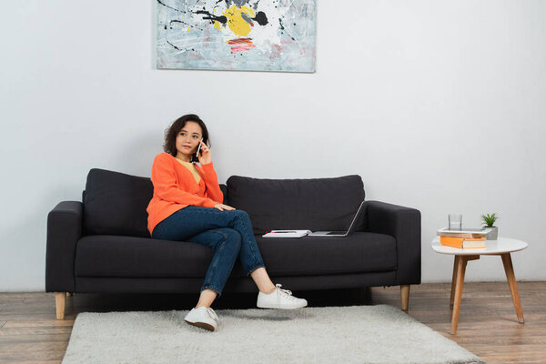 young freelancer talking on smartphone near laptop on couch