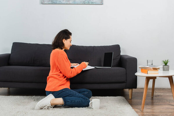 woman sitting on carpet and writing in notebook near laptop with blank screen on couch