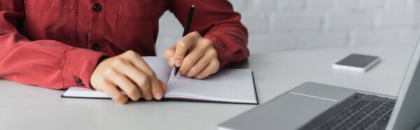 cropped view of teacher writing in notebook near devices on desk, banner