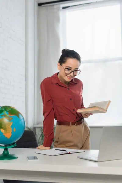 Young Cheerful Teacher Eyeglasses Holding Book Looking Laptop Smartphone Globe — Stock Photo, Image