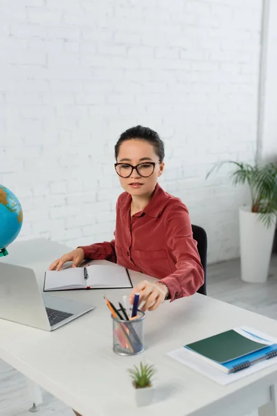 Gözlüklü Genç Öğretmen Masadaki Laptopun Yanındaki Kırtasiyeye Ulaşıyor — Stok fotoğraf